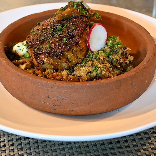 A savory dish in a clay bowl with grilled meat, quinoa, and a garnish of herbs and radish, placed on a white plate.