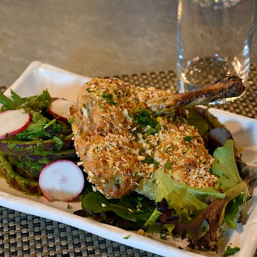 A plated dish consisting of breaded chicken pieces on a bed of mixed greens with sliced radishes, accompanied by an empty glass in the background.