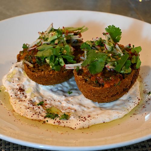 The image shows two falafel patties topped with salad and herbs, placed on a dollop of white sauce on a white plate, garnished with spices.