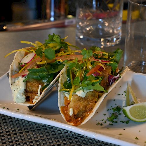 The image shows two fish tacos topped with fresh greens and drizzled sauce on a white rectangular plate with two lime wedges, next to a glass.