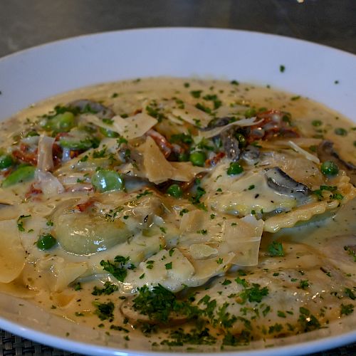 A plate of ravioli in a creamy sauce, garnished with chopped herbs, shaved cheese, mushrooms, and peas.