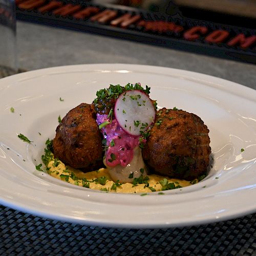 The image shows a white plate with two fried croquettes, topped with herbs, a pink sauce, and a radish slice, served over a yellow creamy base.