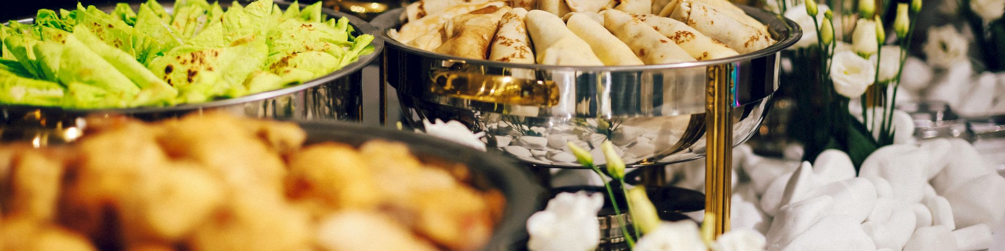 The image shows a buffet setup with various dishes in metal containers, including spring rolls and leafy greens, surrounded by white decorations.